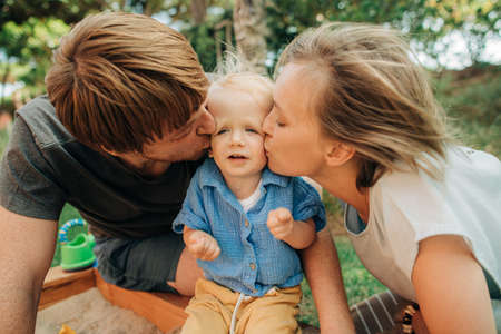 Funny little girl being kissed by parents outdoors. Portrait of cute toddler child sitting confused...