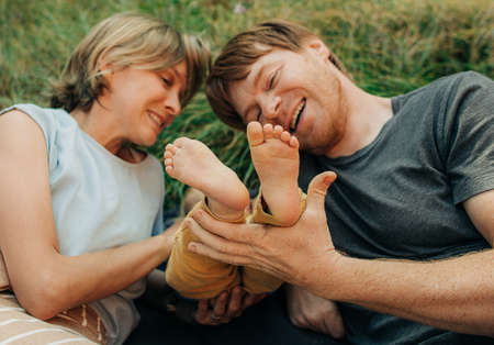 Laughing parents holding feet of their child while lying together outdoors. Family resting on grass...