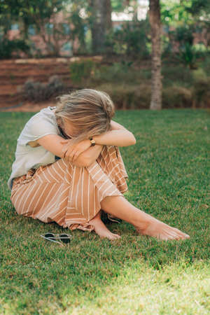 Portrait of upset or tired woman sitting outdoors. Mid adult lady resting on grass with head on arms. Emotional stress conceptの写真素材