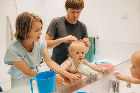 Parents washing their little daughter in bathroom. Cute toddler looking at mirror while washing...