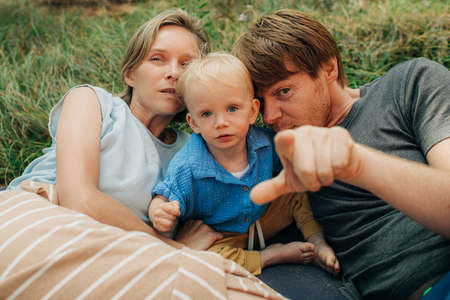 Portrait of happy family lying together on grass. Parents with little child resting outdoors, man showing at camera. Family vacation conceptの写真素材