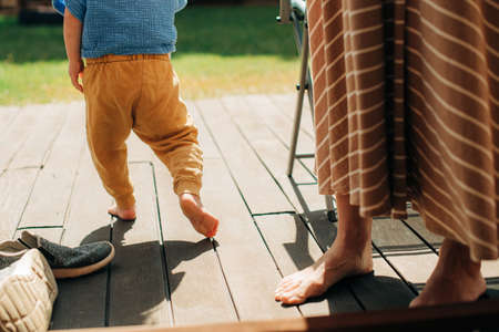 Little child walking on veranda with mother. Close-up of bare legs of woman and toddler standing on terrace in summer. Childcare conceptの写真素材