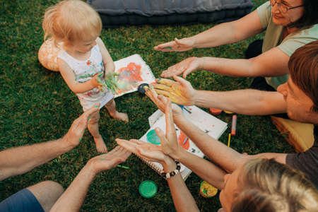 Joyful family reaching hands in paints to toddler. Messy little girl standing on grass at her parents and grandparents among paints and papers. Creativity conceptの写真素材
