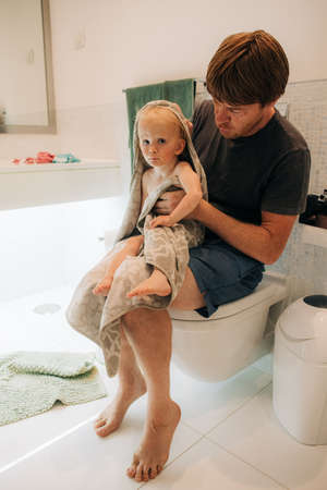 Man sitting on bidets and wrapping baby in towel. Portrait of father drying his daughter in bathroom. Childcare and hygiene conceptの写真素材