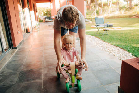 Cute little girl driving toy car with father near their house outdoors. Mid adult man staying with his child at home and playing with her. Father at home, childhood conceptの写真素材