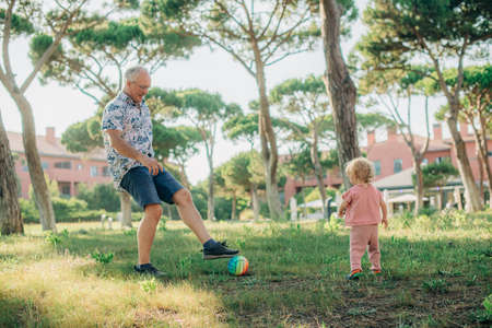 Happy grandfather playing football with toddler in park. Little granddaughter walking with grandpa...