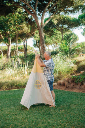 Mature man adjusting textile wigwam tent outdoors. Portrait of grandfather putting tent in backyard....