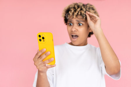 Portrait of shocked African American woman. Female model in glasses with curly hair looking at mobile phone with shock. Portrait, studio shot, emotion conceptの写真素材