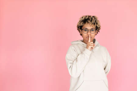 Serious woman in glasses calling be silent. African American female model in hoodie touching lips...