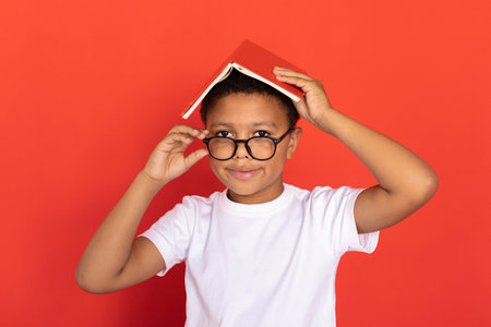 Portrait of happy schoolboy standing with book on head and looking over glasses. Mixed race preteen...