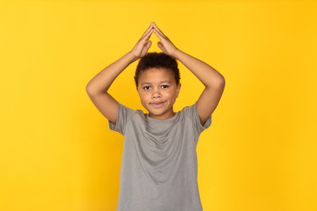 Portrait of happy preteen boy making roof gesture over head. Mixed race child wearing gray T-shirt looking at camera and smiling against yellow background. Childs security, insurance conceptの写真素材