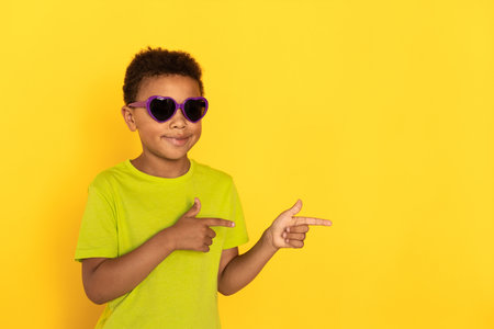 Portrait of happy preteen boy in sunglasses pointing direction. Mixed race child wearing green T-shirt showing something against yellow background. Presentation conceptの写真素材