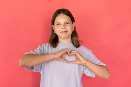 Portrait of happy preteen girl making heart gesture. Caucasian child wearing blue T-shirt showing love symbol against red background. Love conceptの写真素材