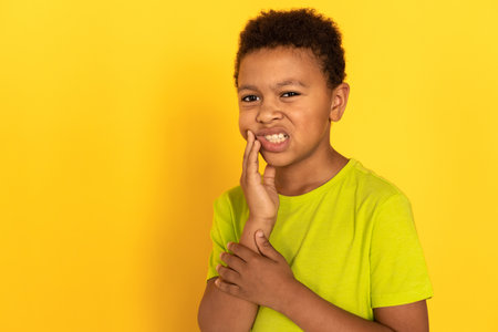 Portrait of sad preteen boy suffering from toothache. Mixed race child wearing green T-shirt touching aching cheek against yellow background. Childs health problems conceptの写真素材