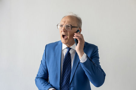 Portrait of angry mature businessman talking to client on phone. Senior manager wearing formalwear and eyeglasses shouting in his smartphone against white background. Angry boss conceptの写真素材