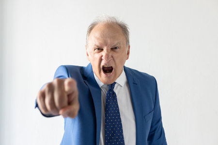 Portrait of furious mature businessman pointing at camera. Senior manager wearing formalwear looking at camera and shouting against white background. Angry boss conceptの写真素材