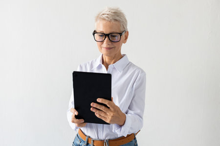 Portrait of senior woman working on tablet. Female model in white shirt holding tablet, working. Portrait, studio shot, technology conceptの写真素材