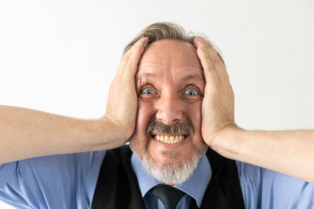 Close-up of excited or frustrated mature businessman. Senior Caucasian manager wearing formalwear staring, clenching teeth and holding head in hands against white background. Shock or awe conceptの写真素材