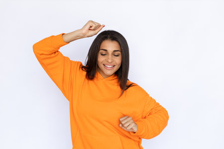 Portrait of joyful Hispanic woman dancing. Happy young model with long hair in orange hoodie celebrating success or having good time with closed eyes. Happiness, entertainment concept.の写真素材