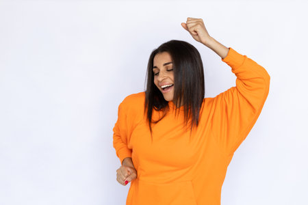 Portrait of happy Hispanic woman dancing and singing. Excited young model with long hair in orange...