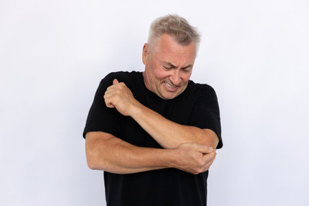 Senior man holding elbow. Male model in black T-shirt experiencing pain in elbow with painful expression. Portrait, studio shot, pain conceptの写真素材