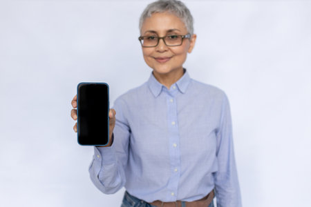 Portrait of positive senior businesswoman showing smartphone. Mature woman wearing eyeglasses and blue shirt advertising new mobile phone. Mobile technology conceptの写真素材
