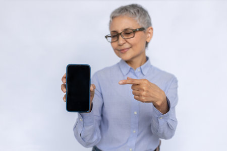 Portrait of positive senior businesswoman pointing at smartphone. Mature woman wearing eyeglasses and blue shirt presenting new model of mobile phone. Mobile technology conceptの写真素材