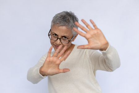Portrait of confident senior woman protecting herself with hands. Scared mature Caucasian woman wearing eyeglasses and white jumper making forbiddance gesture. Fear and forbiddance conceptの写真素材