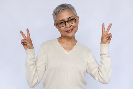 Portrait of happy senior woman in glasses making peace gesture. Mature Caucasian woman wearing white jumper looking at camera and smiling over white background. Happiness conceptの写真素材