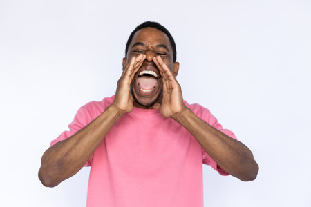 Portrait of loud African American man shouting. Frowned young male model with short dark hair in pink T-shirt with closed eyes screaming with hands near mouth. Studio shot, announcement conceptの写真素材