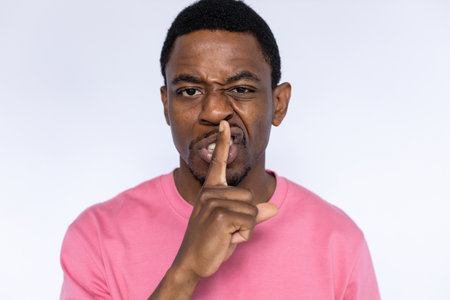 Portrait of angry African American man shushing. Displeased young male model with short dark hair in pink T-shirt looking at camera with finger on lips, asking to be quiet. Secret, silence conceptの写真素材