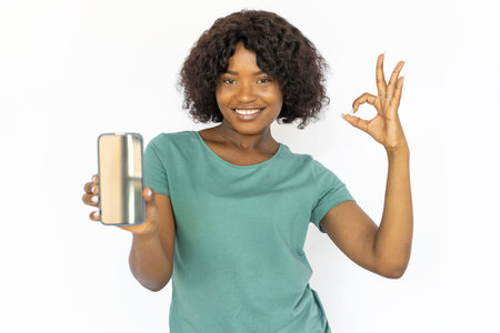 Good-looking young woman with smartphone showing OK sign. Happy African American girl demonstrating smartphone screen, making OK gesture. Studio shoot, technology, advertisement conceptの写真素材