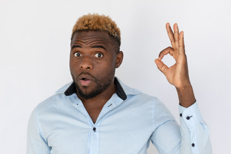 Portrait of shocked young man with ok gesture. Amazed young male model with blond hair in light blue shirt looking at camera, recommending product. Advertisement, approval conceptの写真素材