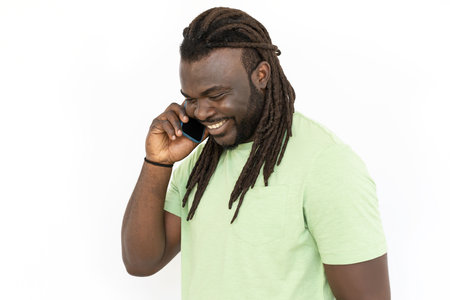 African American man with dreadlocks talking on phone, smiling. Black man standing against white background, looking down, having conversation with friend. Communication, happiness conceptの写真素材