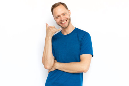 Happy man in T-shirt making phone call gesture. Cheerful Caucasian man showing call me gesture at camera while standing on white background. Communication, conversation conceptの写真素材