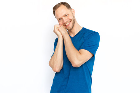 Touched man smiling at camera, holding hands together over cheek. Man being kind, affected or melting from cuteness while standing on white background. Trust, kindness, love, appreciation conceptの写真素材