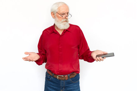 Unsure senior man with phone shrugging. Upset Caucasian male model with gray hair and beard in red shirt and glasses looking at screen with helpless gesture. Modern technology, advertising conceptの写真素材