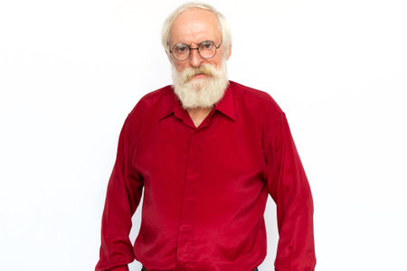 Angry senior man frowning. Portrait of displeased Caucasian male model with gray hair and beard in red shirt and glasses looking at camera, arguing or reprimanding someone. Conflict, anger conceptの写真素材