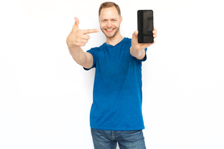 Happy man pointing at blank phone screen with index finger. Smiling man looking at camera, promoting product while standing on white background. Advertisement, promotion conceptの写真素材