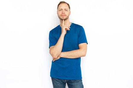 Pensive man looking at camera, touching chin with finger. Peaceful man in T-shirt looking at camera while standing on white background. Imagination, wonder, inspiration conceptの写真素材