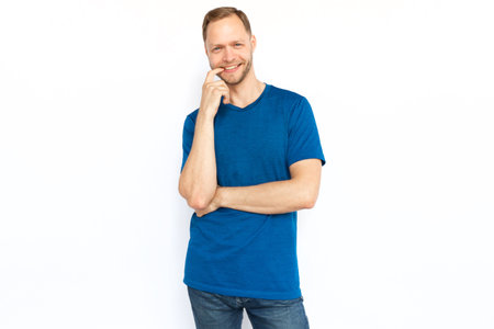 Happy man smiling at camera on white background. Caucasian guy with fair hair in T-shirt looking and posing for camera with hand on his mouth. Happiness, positivity conceptの写真素材