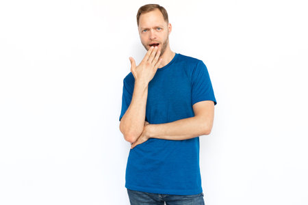Shocked man covering his mouth with hand. Bearded Caucasian man receiving excited news while standing on white background and looking at camera. Shock, secret, amazementの写真素材