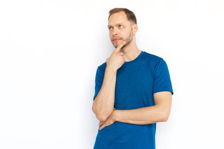 Pensive Caucasian man thinking and looking up. Concentrated guy touching chin with finger, considering ideas or dreaming while standing on white background. Imagination, wonder, inspiration conceptの写真素材