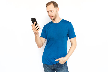 Shocked man looking at mobile phone screen. Surprised bearded man receiving bad or good news, standing on white background, posing for camera. Emotion, surprise, frustration conceptの写真素材