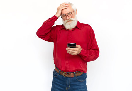 Disappointed senior man using phone with hand on head. Tired Caucasian male model with gray hair and beard in red shirt and glasses looking at screen, covering forehead. Modern technology conceptの写真素材