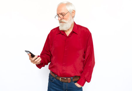 Happy senior man using phone. Pleased Caucasian male model with gray hair and beard in red shirt and glasses looking at screen with hand in pocket, watching video. Modern technology conceptの写真素材