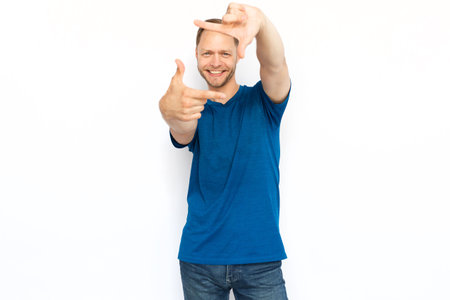 Happy man showing frame gesture at camera. Smiling Caucasian man looking at camera through frame formed by his hands while standing on white background. Imagination, creativity conceptの写真素材