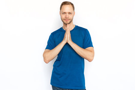 Peaceful man with praying hands looking at camera. Cheerful man in T-shirt looking at camera while standing on white background, praying, making wish or meditating.Harmony, hope, concentration conceptの写真素材