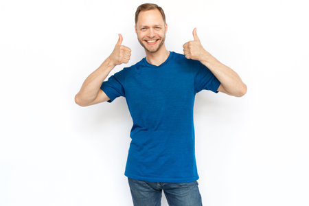 White man showing thumbs up while standing on white background. Happy bearded man in T-shirt looking at camera and showing approving gestures. Agreement, approval conceptの写真素材