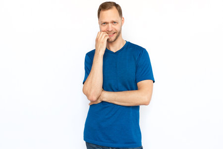 Nervous man biting his nails while standing on white background. Concerned Caucasian man looking at camera, feeling stressed and worried. Stress, anxiety, fear conceptの写真素材